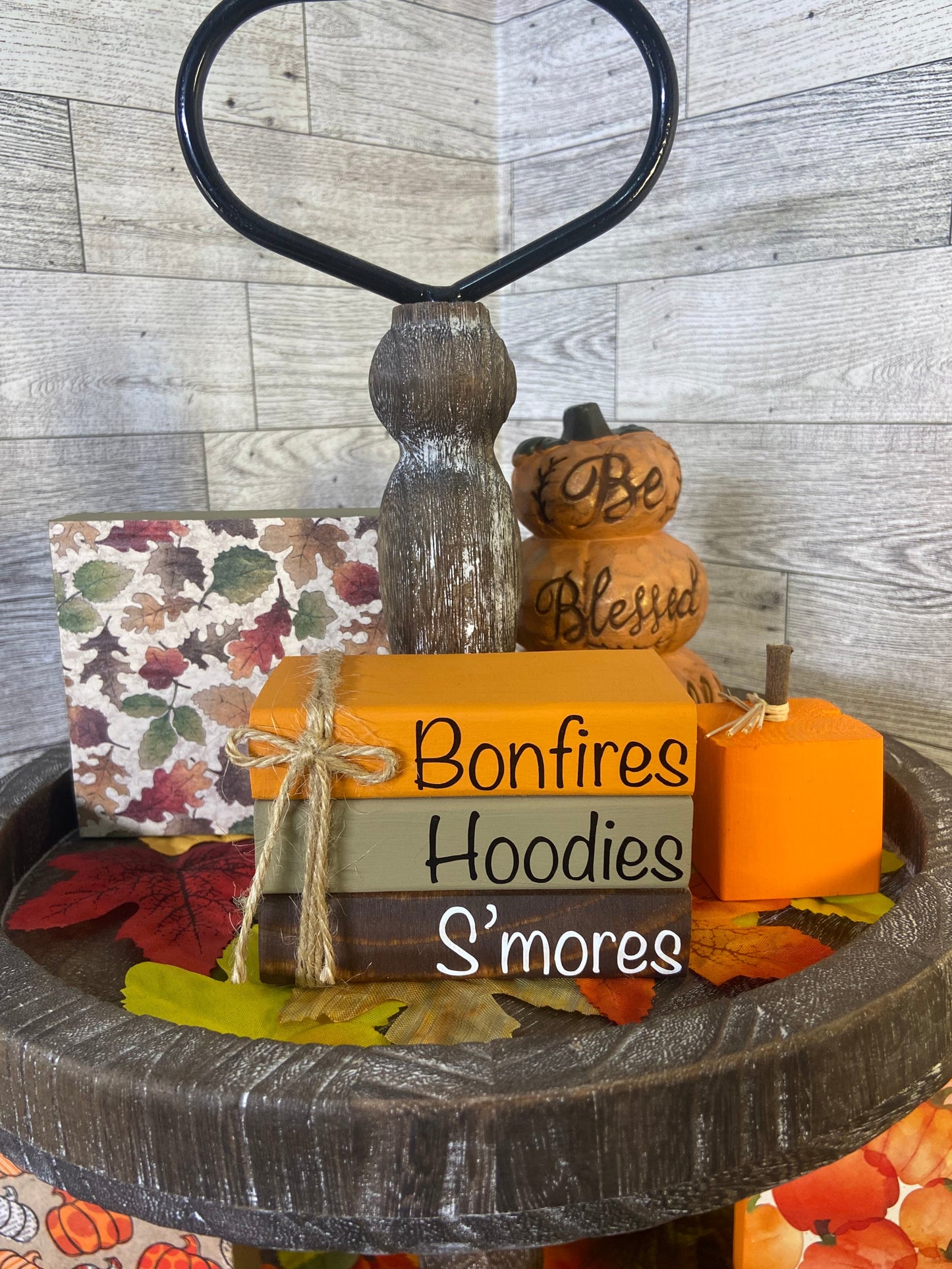 A handcrafted wooden book stack with fall colors and 'Bonfires Hoodies S'mores' text, adorned with jute rope and vinyl lettering, displayed on a tiered tray with autumn leaves around it.